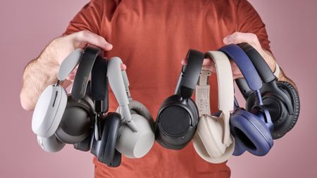 A man holding eight pairs of headphones in his hands, with models from Bose, Sony, Amazon, Soundcore, and Sonos, among others